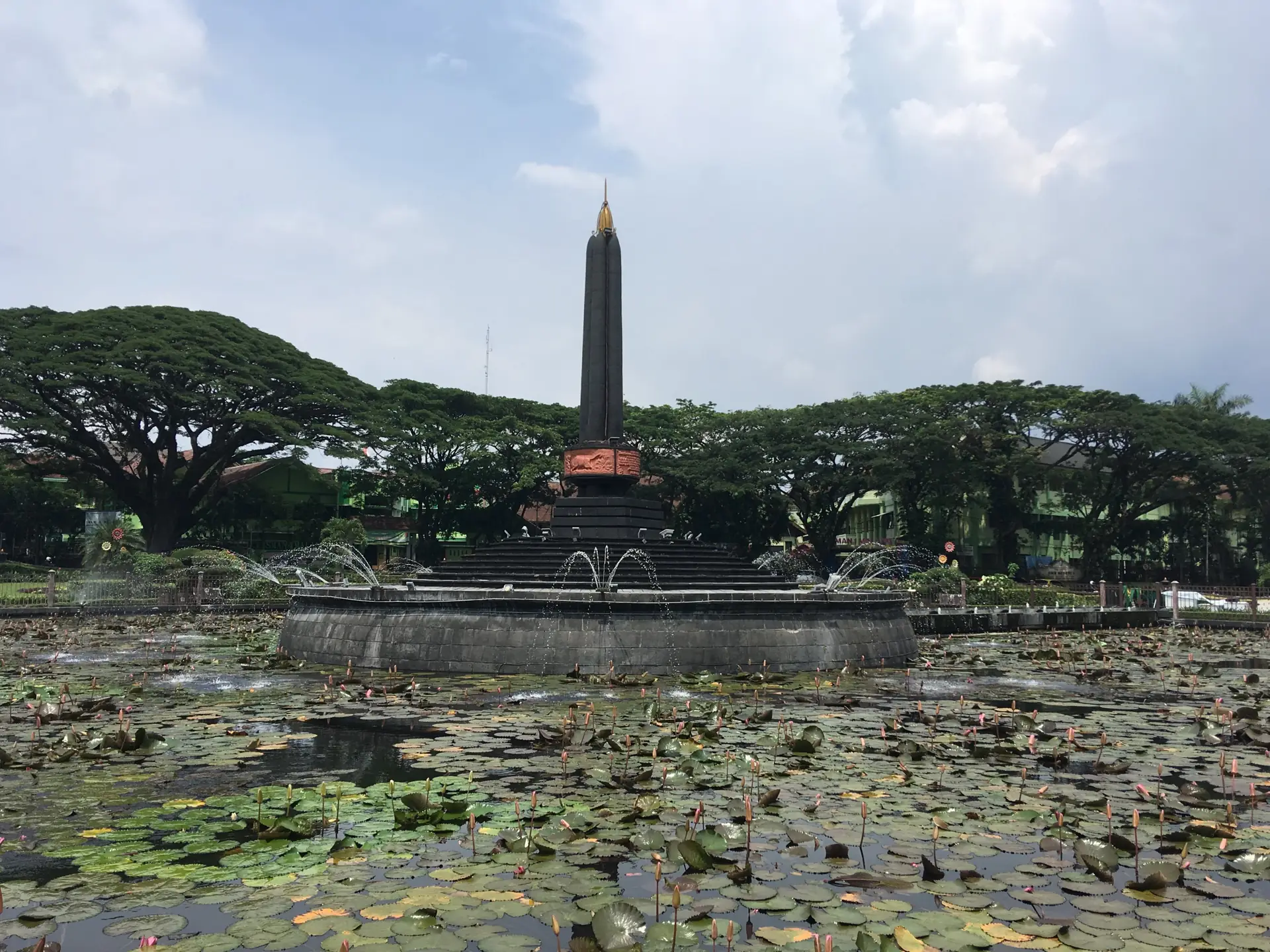 Monumen Tugu Malang berbentuk obelisk hitam di tengah kolam teratai dan air mancur Alun-Alun Tugu, dikelilingi pohon trembesi besar pada siang hari