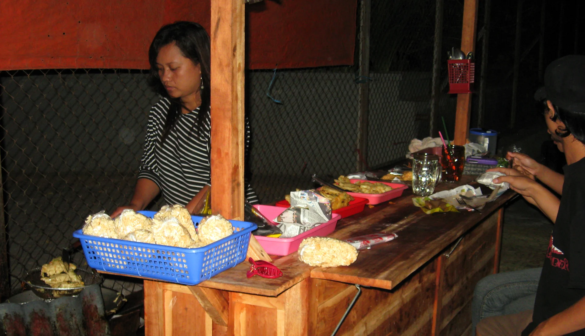 Penjual angkringan pada malam hari dengan bungkusan nasi kucing di wadah merah muda, keranjang kerupuk, dan pembeli makan di meja kayu