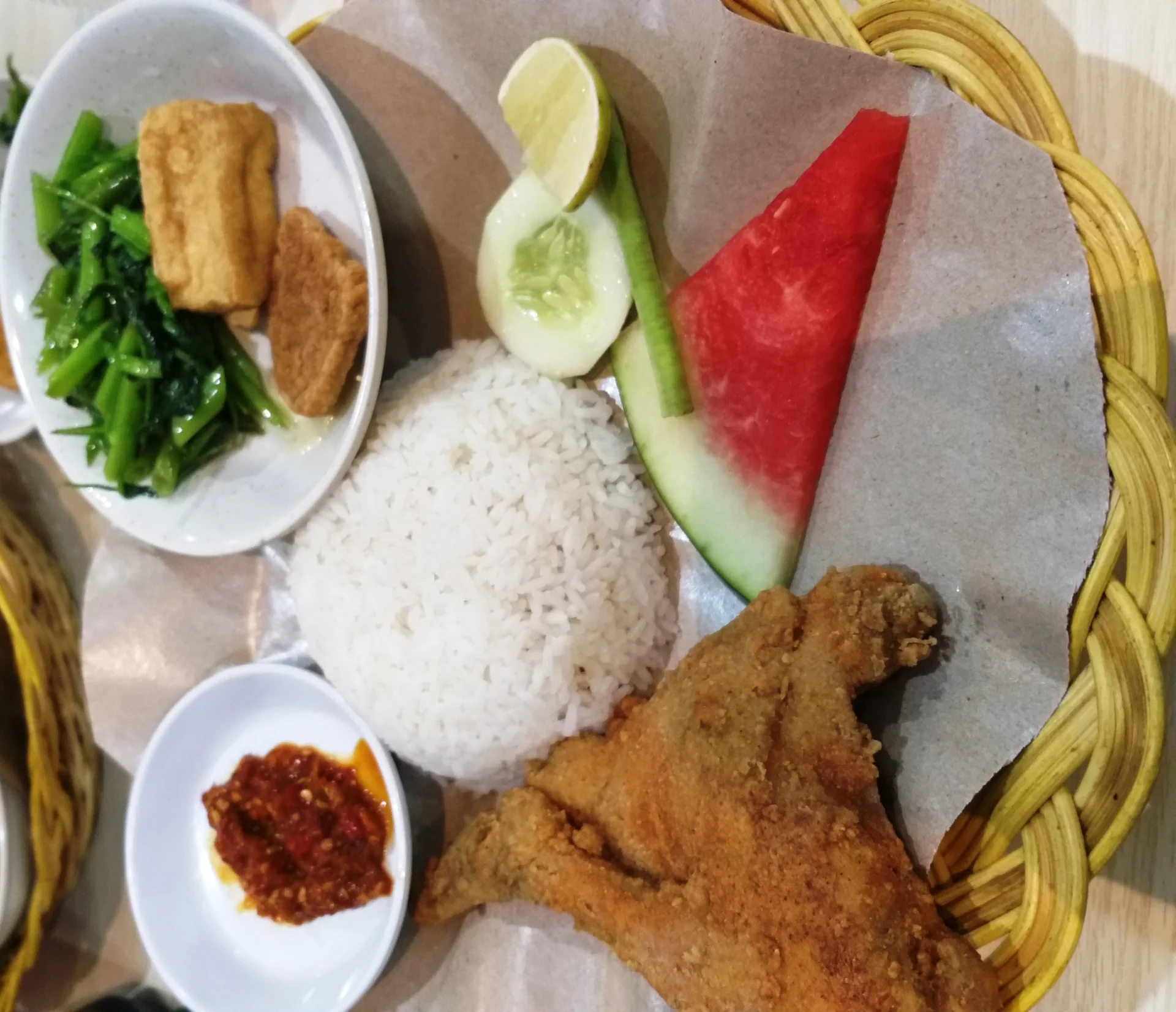 paket ayam goreng tepung dengan nasi, tumis kangkung, tahu tempe, sambal, timun, dan semangka di keranjang rotan
