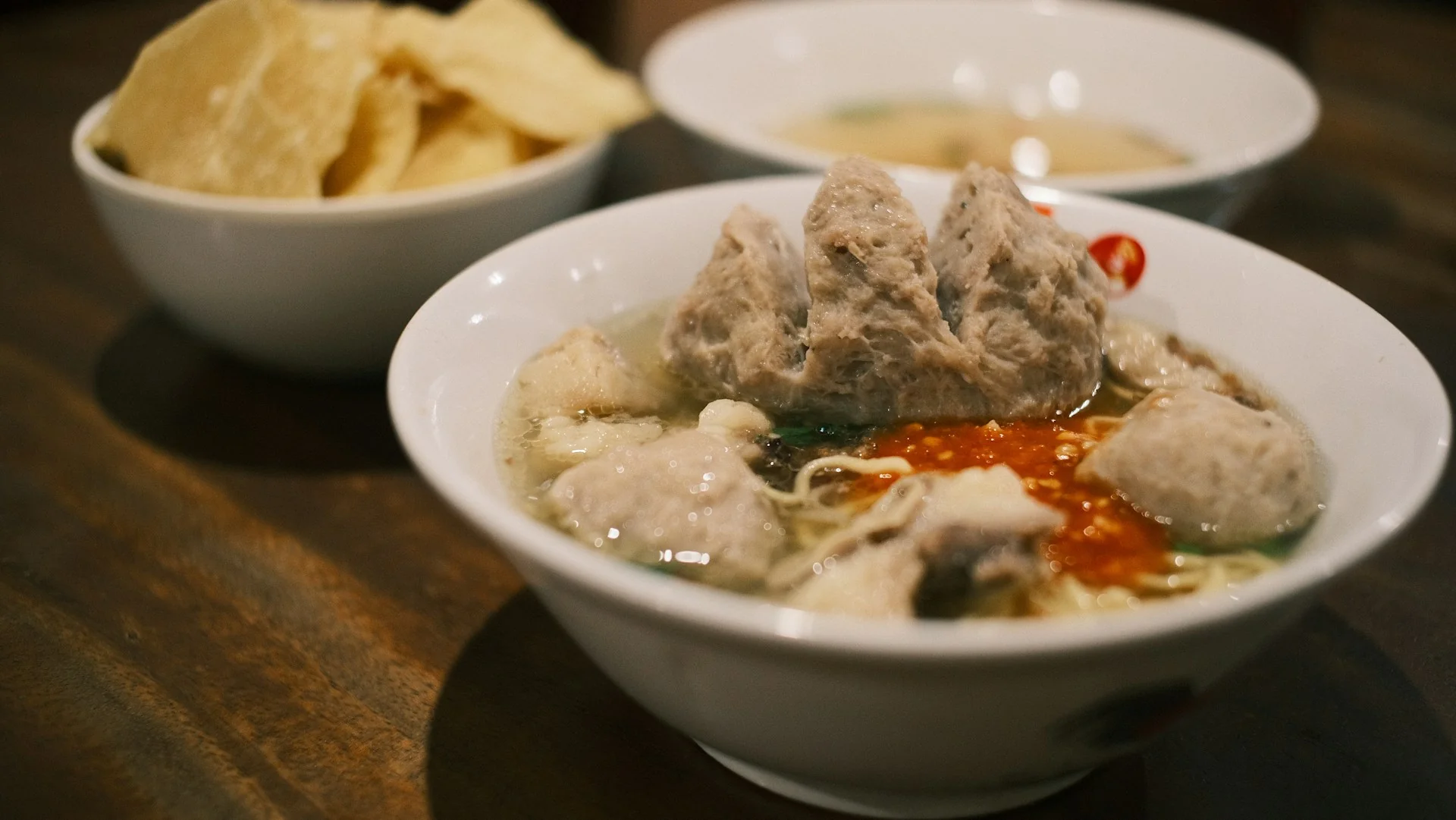 semangkuk bakso kuah dengan bakso besar, mi kuning, dan sambal, ditemani mangkuk kerupuk di meja kayu