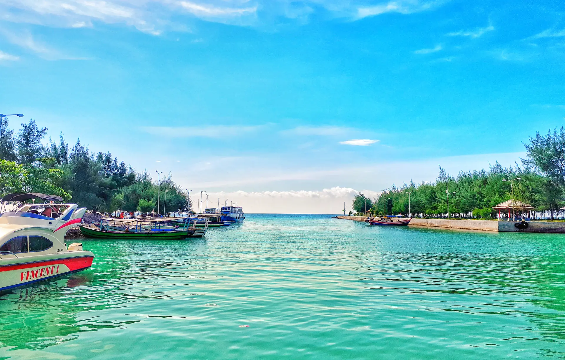 Perairan hijau toska di dermaga Pulau Pari dengan speedboat dan perahu kayu bersandar, diapit pohon cemara laut