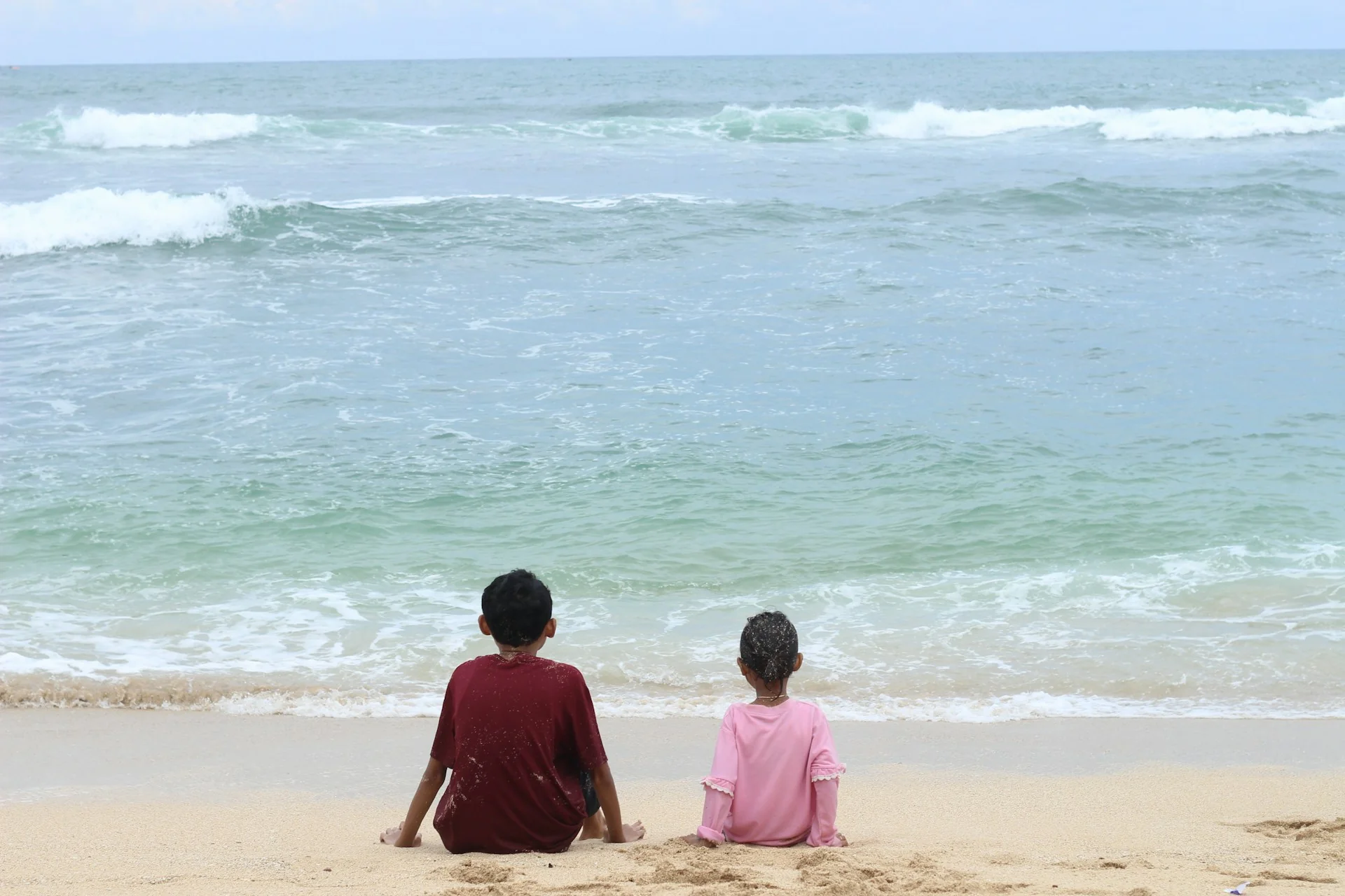 dua anak duduk di pasir pantai membelakangi kamera sambil memandang ombak