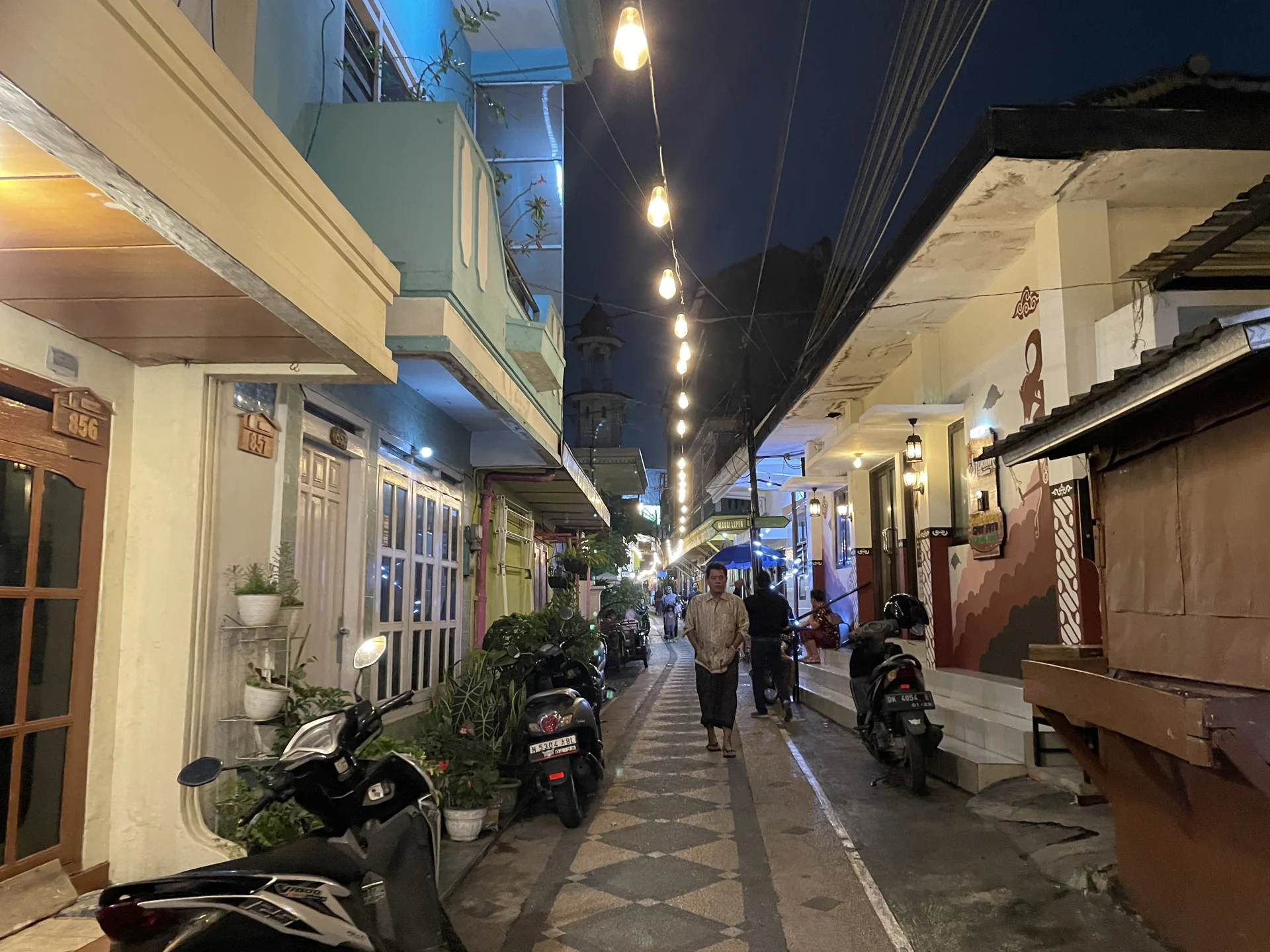 Gang sempit Kampung Heritage Kayutangan, Malang, pada malam hari dengan untaian lampu bohlam, rumah-rumah lama berwarna pastel, sepeda motor terparkir, dan warga berjalan di jalan berpola