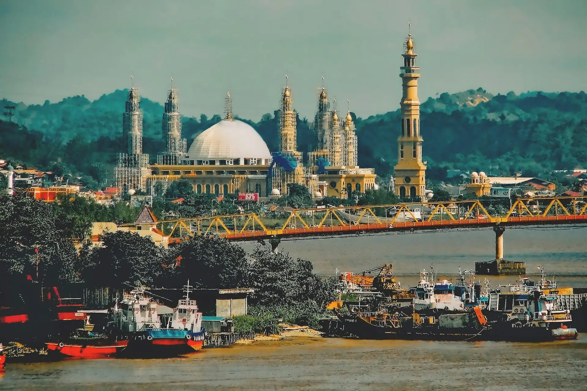 Jembatan Mahakam berwarna kuning merah di atas Sungai Mahakam dengan kapal tunda di tepi dan menara Islamic Center di latar