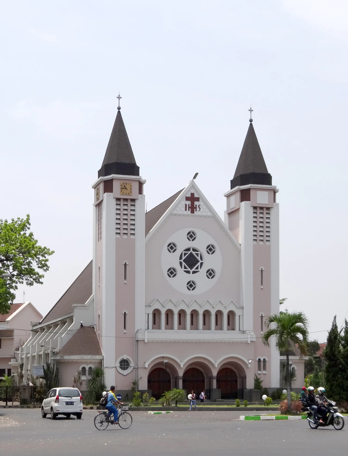 Fasad Gereja Katedral Ijen Malang bercat merah muda pucat dengan dua menara runcing dan jendela mawar, dilihat dari jalan pada siang hari