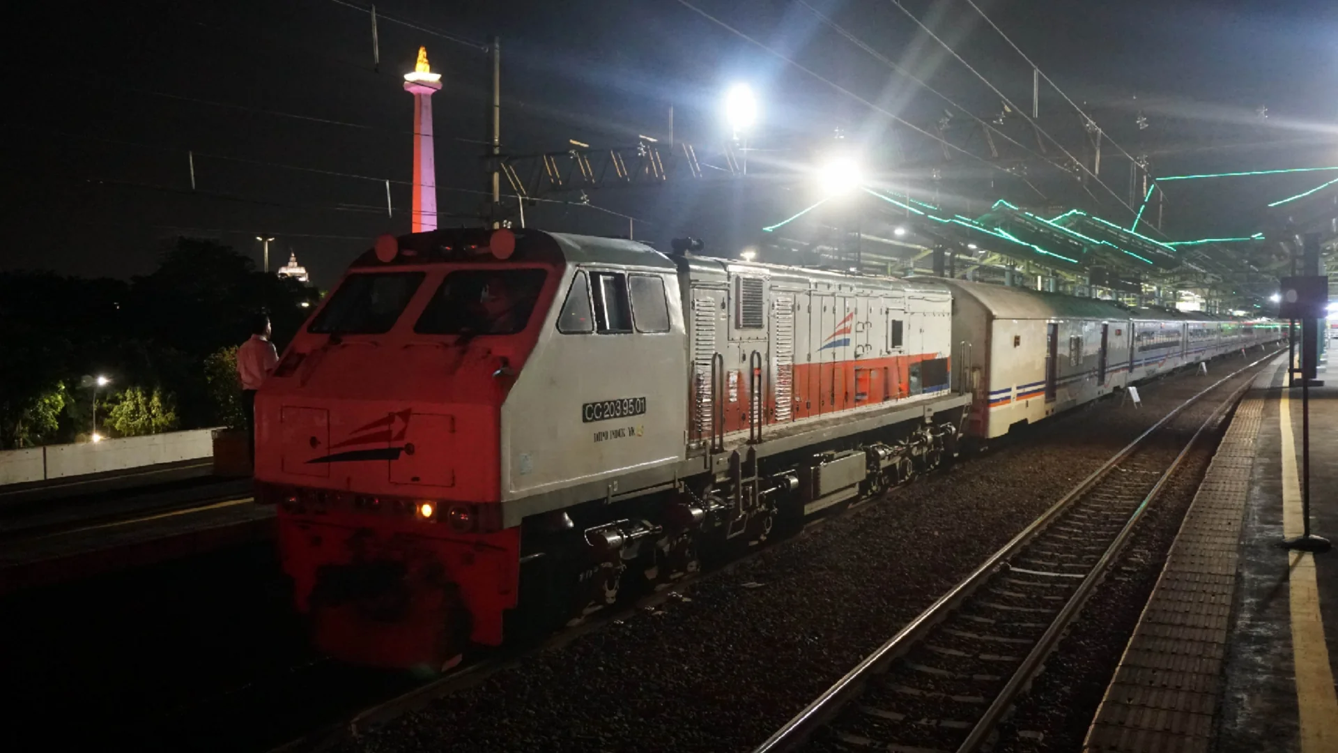 Lokomotif merah-putih menarik rangkaian Kereta Api Sembrani di peron Stasiun Gambir pada malam hari dengan Tugu Monas di latar belakang