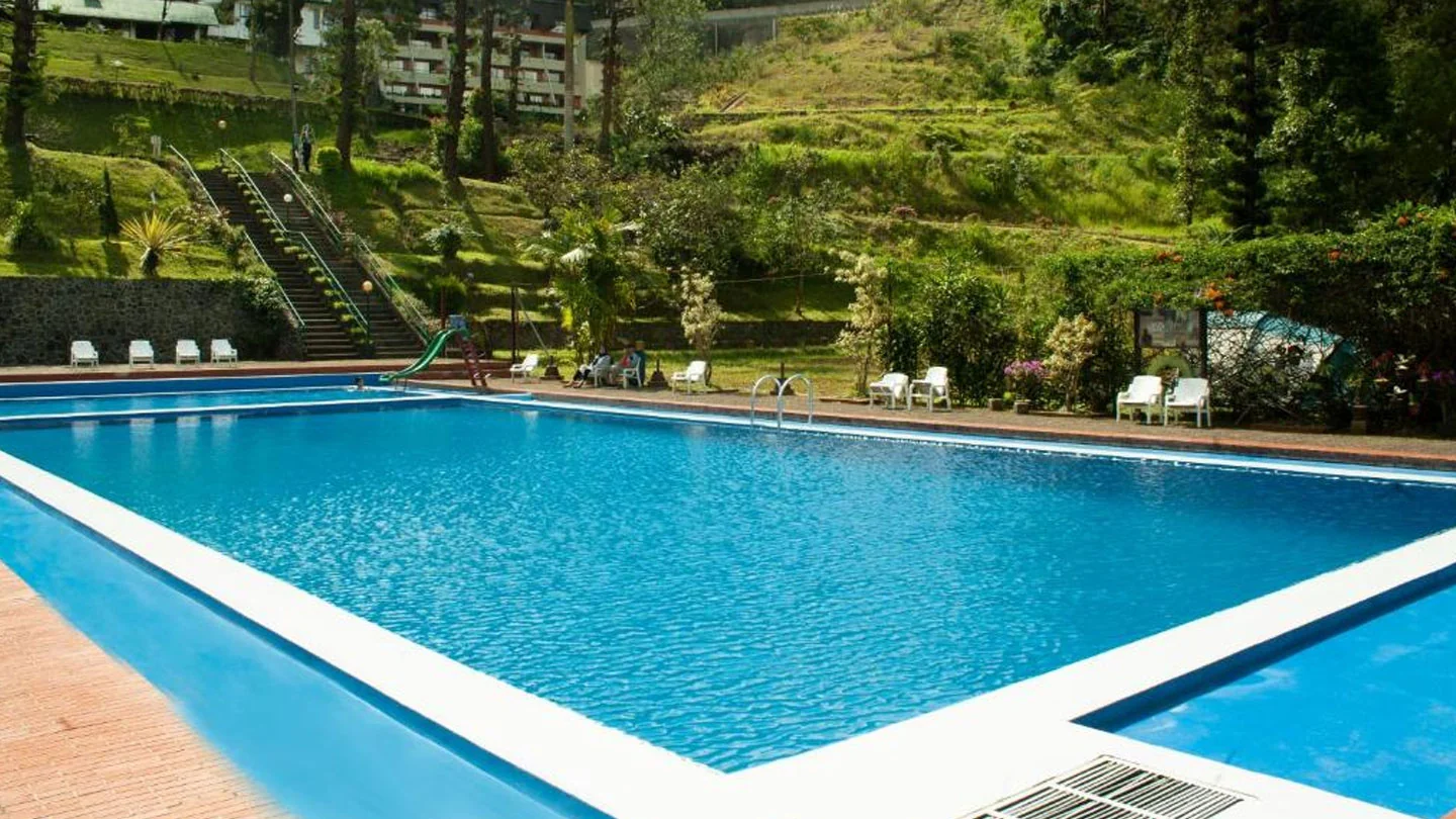 Kolam renang outdoor Puncak Pass Resort berair biru dengan kursi santai putih dan taman di lereng bukit