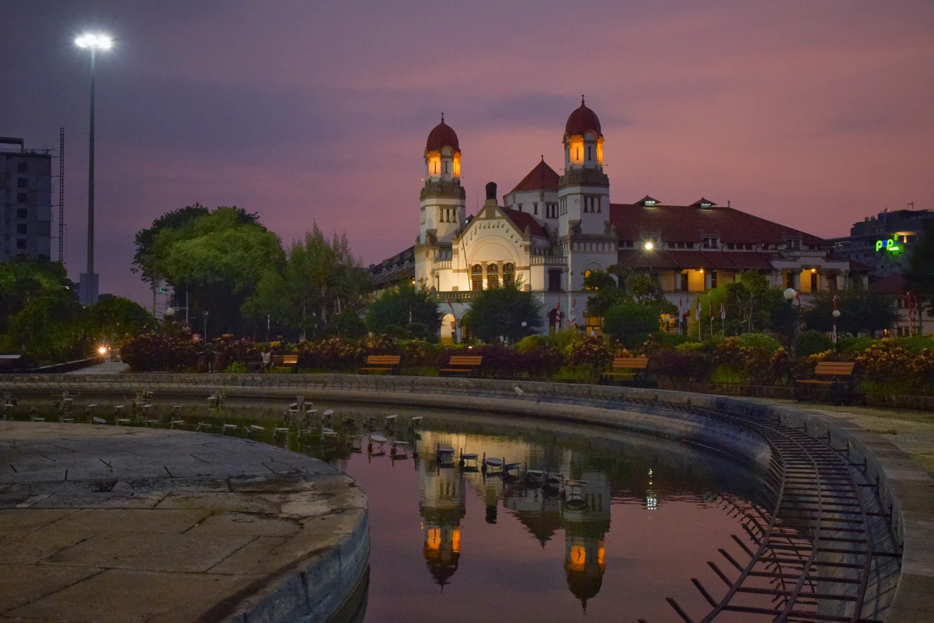Gedung Lawang Sewu Semarang dengan dua menara berlampu di bawah langit ungu menjelang fajar, terpantul di kolam Tugu Muda