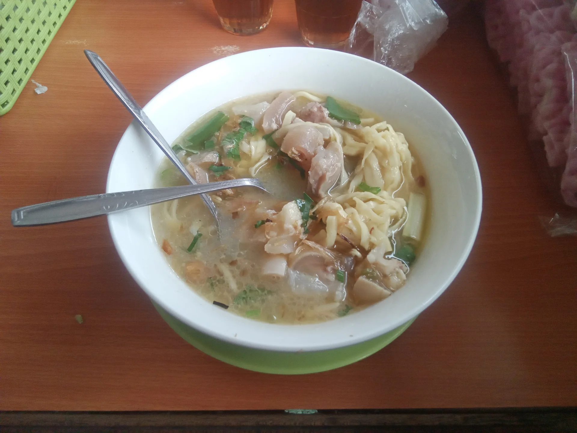 mangkuk putih berisi mie kocok dengan potongan kikil kaki sapi, mi kuning pipih, seledri, dan bawang goreng di meja warung