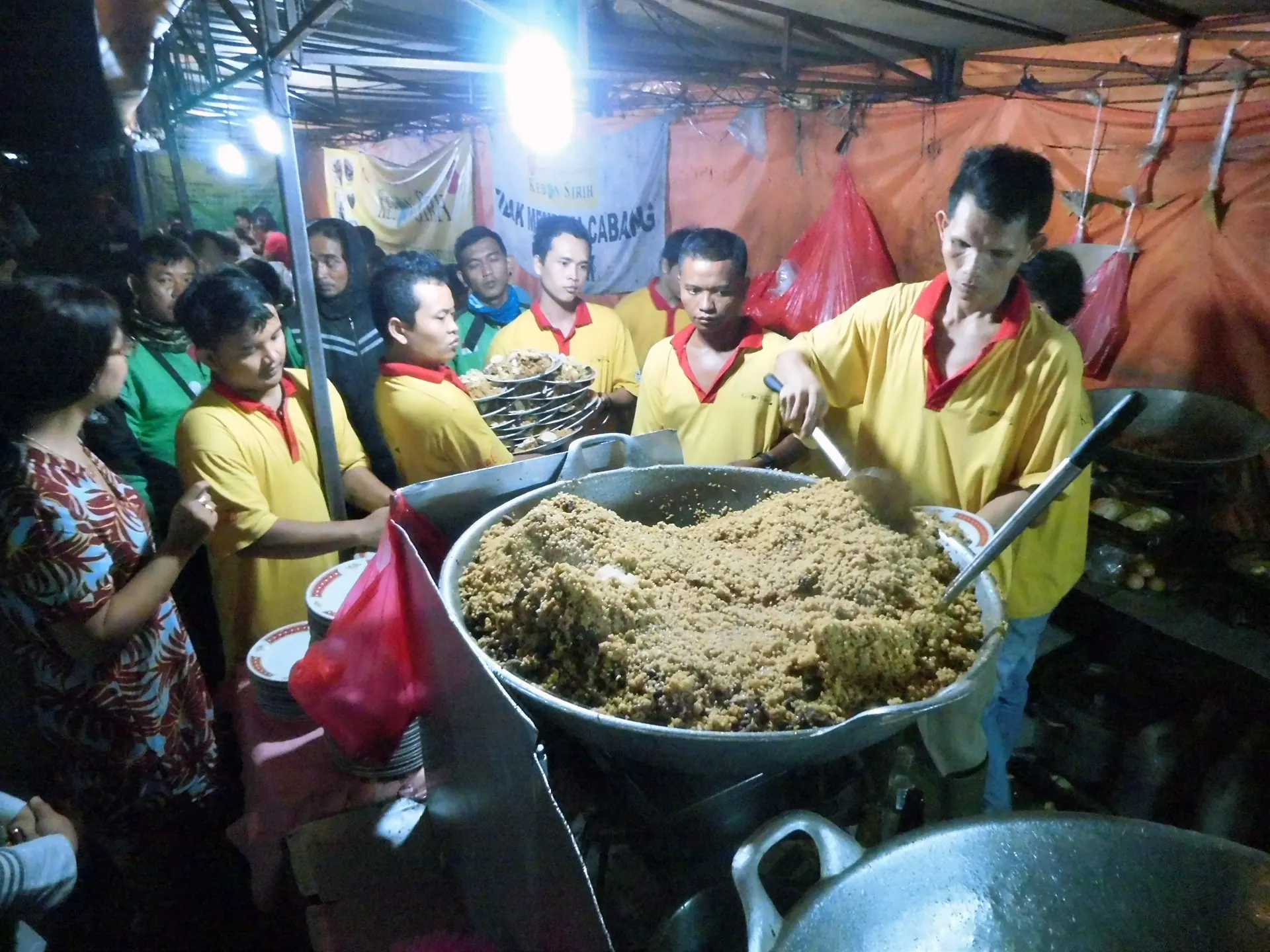 juru masak mengaduk nasi goreng kambing dalam wajan besar di tenda kaki lima malam, dikerumuni pembeli dan pelayan