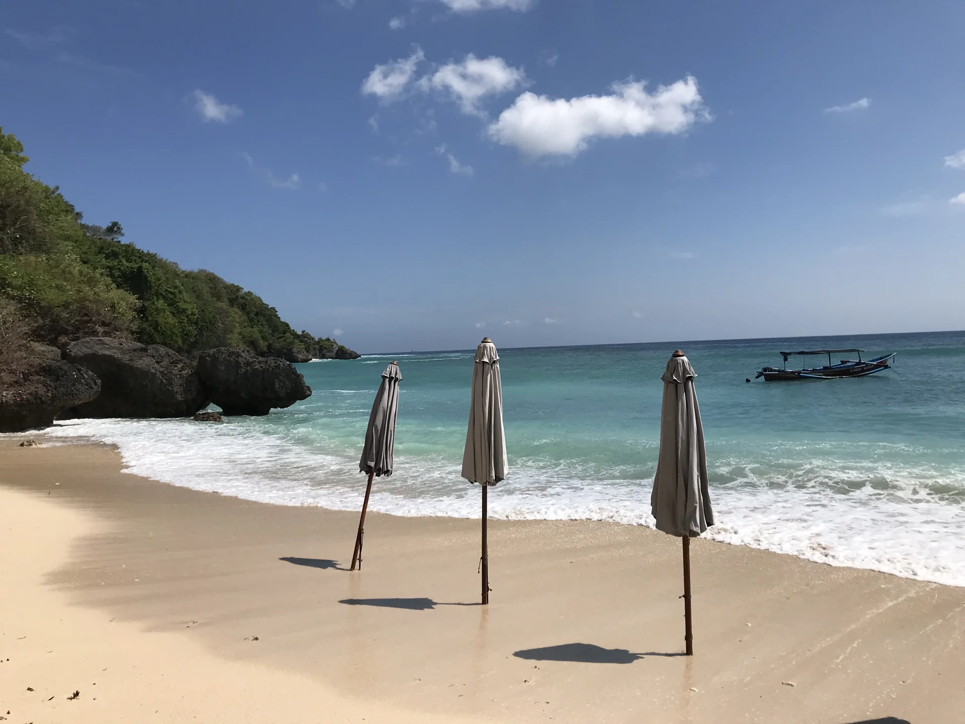 Pasir putih Pantai Padang Padang dengan payung tertutup, perahu nelayan, dan tebing hijau di Pecatu, Bali