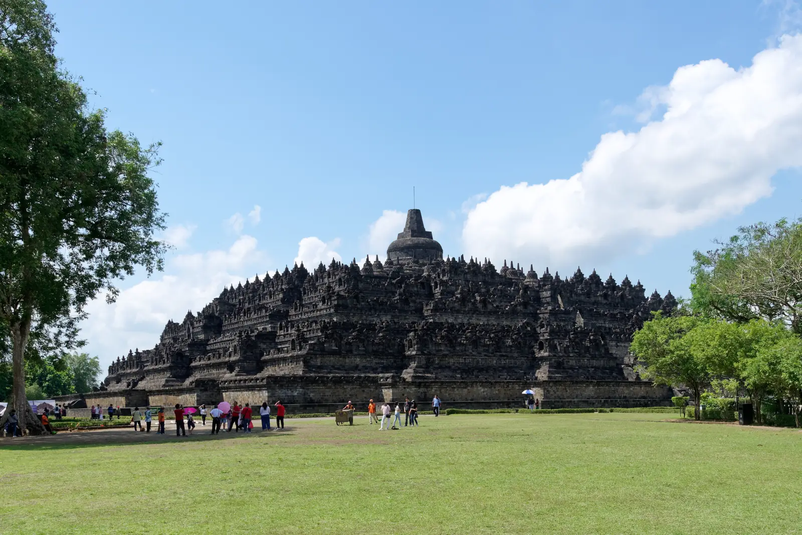 Pelataran rumput luas di depan Candi Borobudur dengan pengunjung berjalan di bawah langit biru