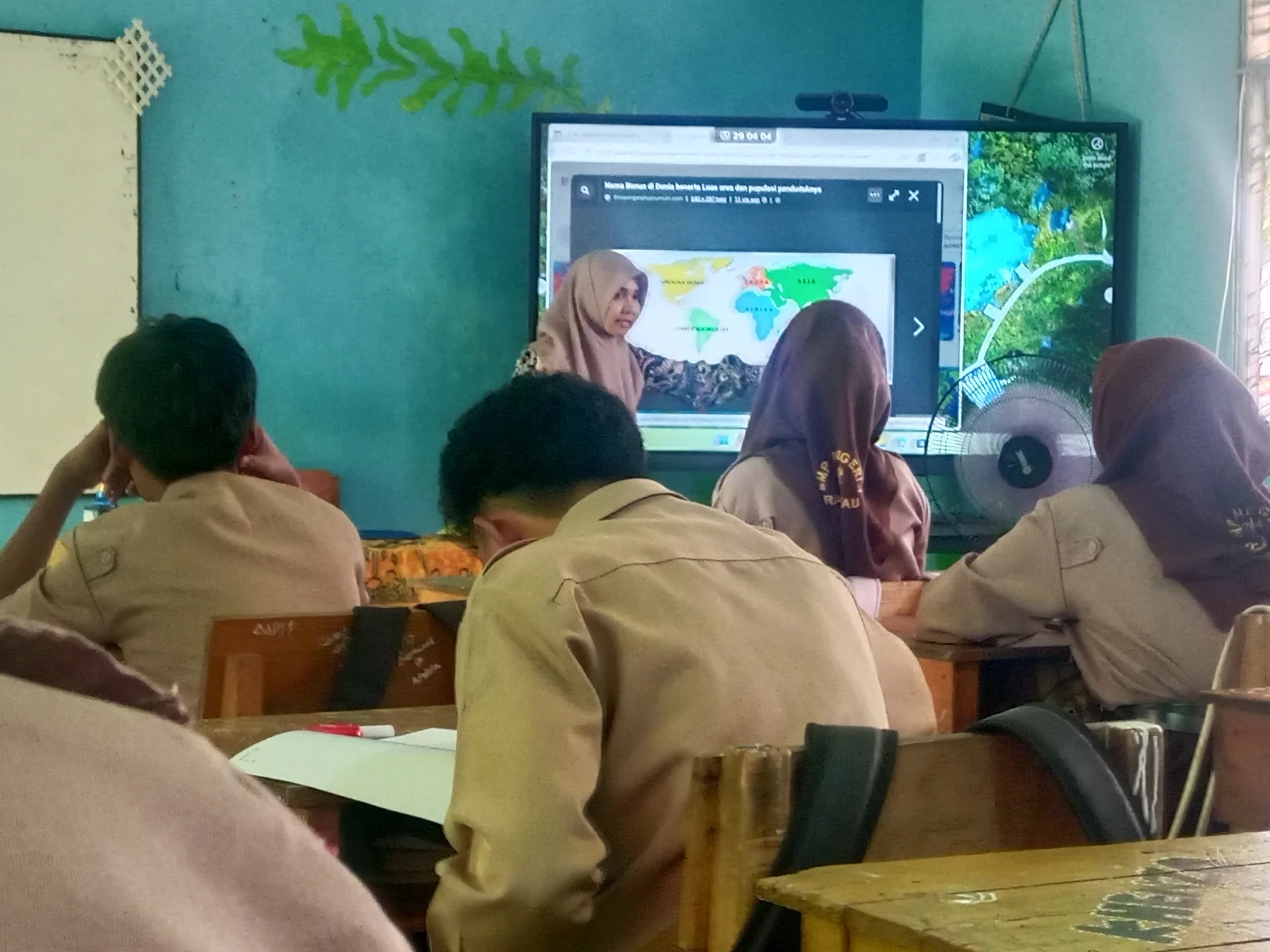 suasana ruang kelas SMP dengan siswa berseragam pramuka memperhatikan guru di depan layar