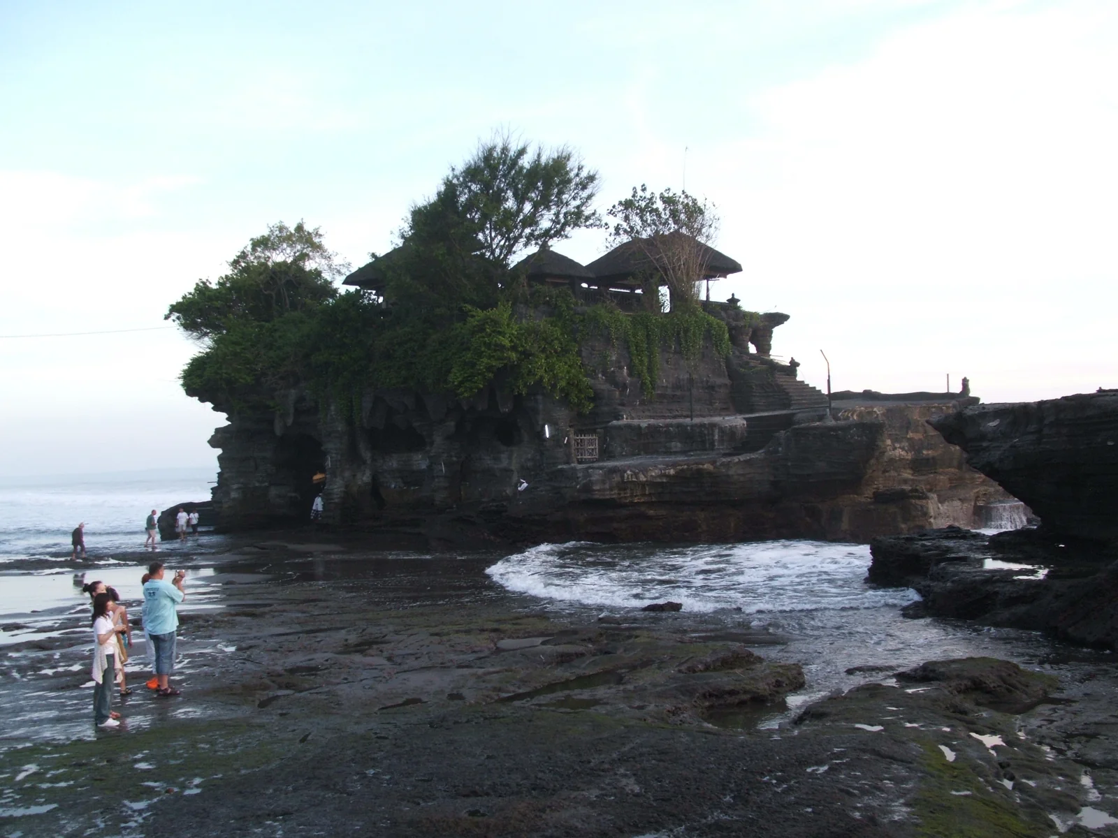 Pengunjung berdiri di dasar karang yang terbuka saat air laut surut di kaki Pura Tanah Lot, Tabanan, Bali