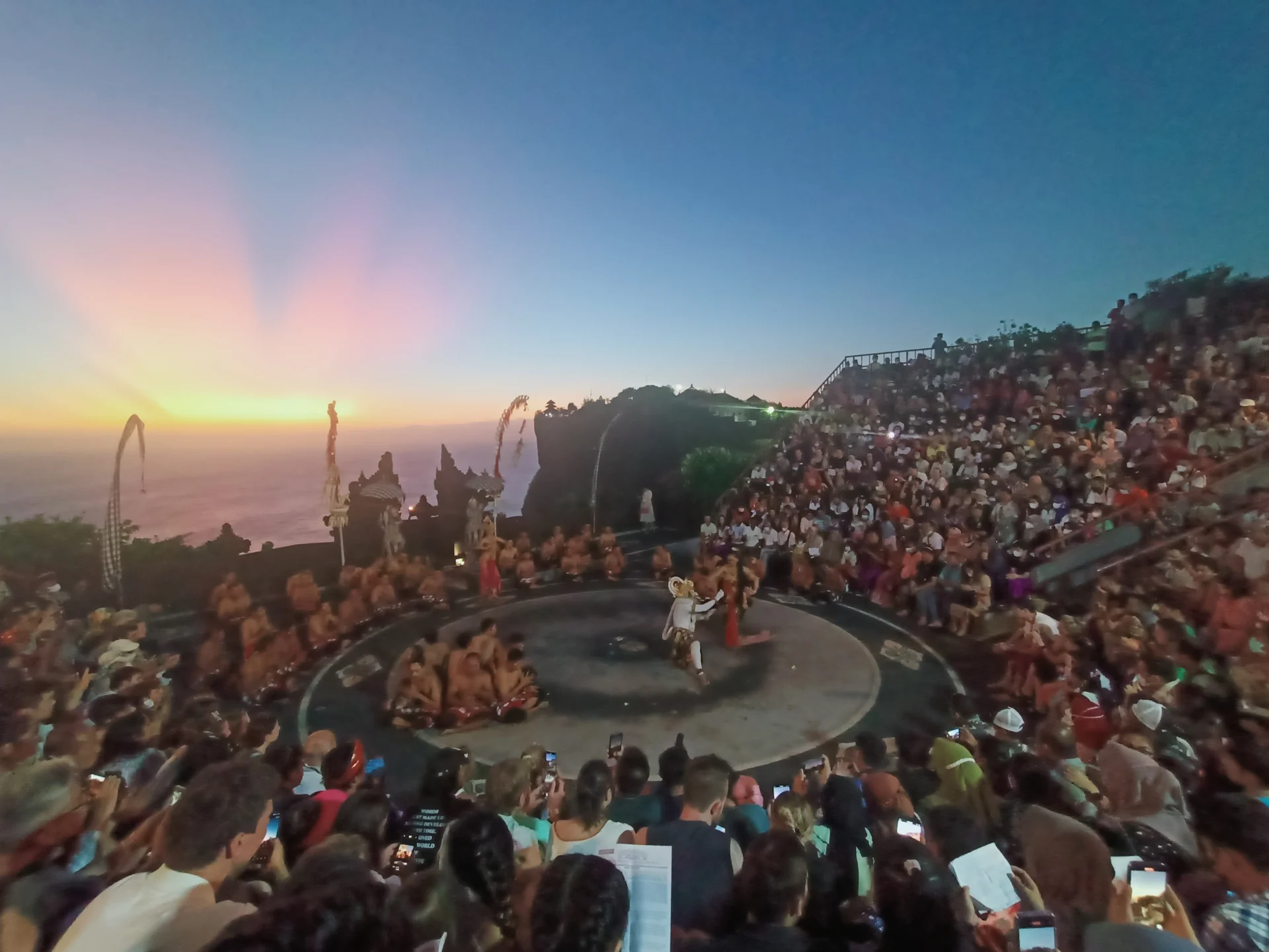 Pertunjukan Tari Kecak di amfiteater Uluwatu saat senja dengan penonton memadati tribun dan laut di latar belakang