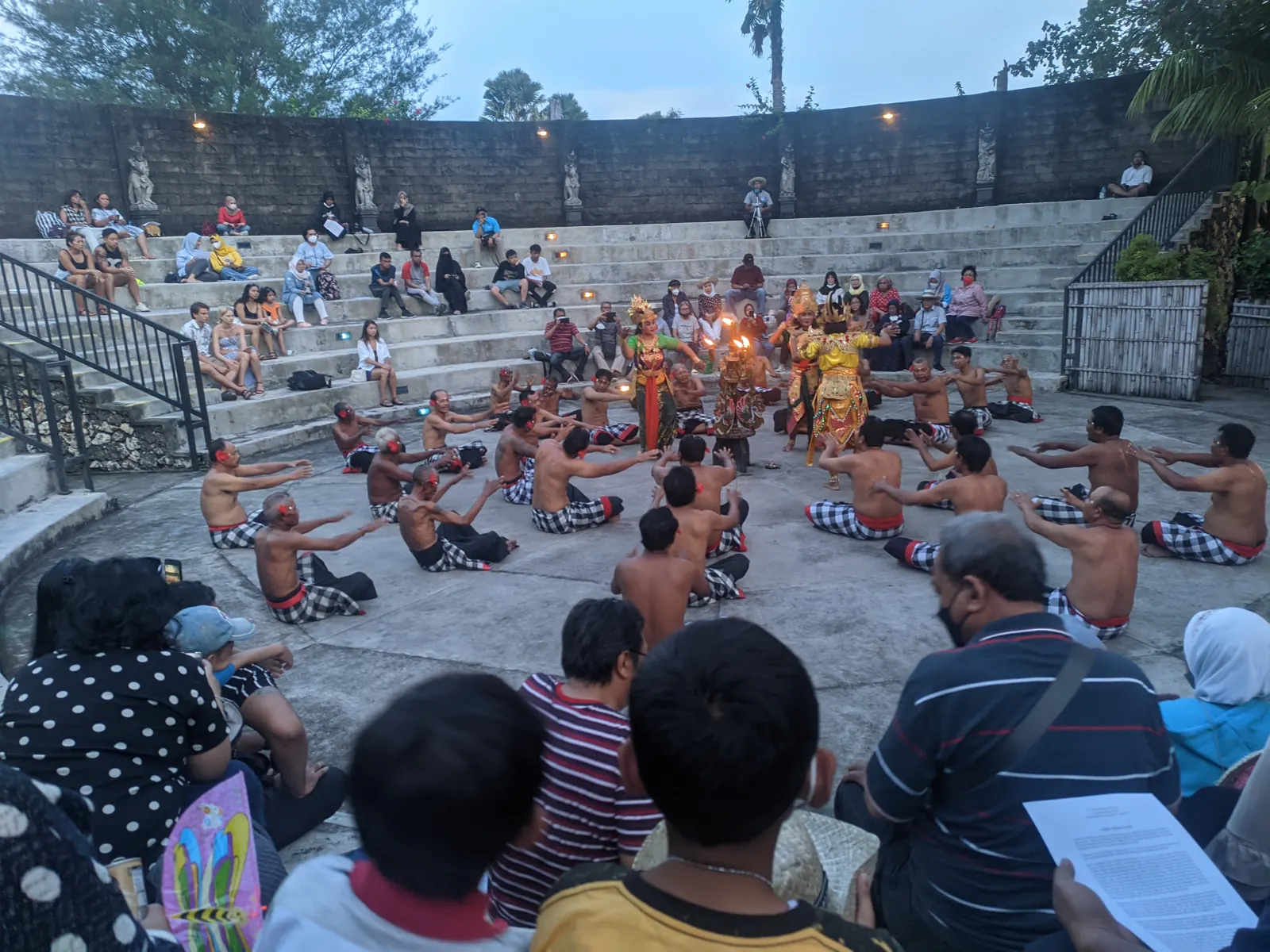 Penari Kecak bertelanjang dada duduk melingkar mengelilingi penari berkostum dan bermahkota di amfiteater terbuka Tanah Lot saat senja, ditonton pengunjung di tribun batu