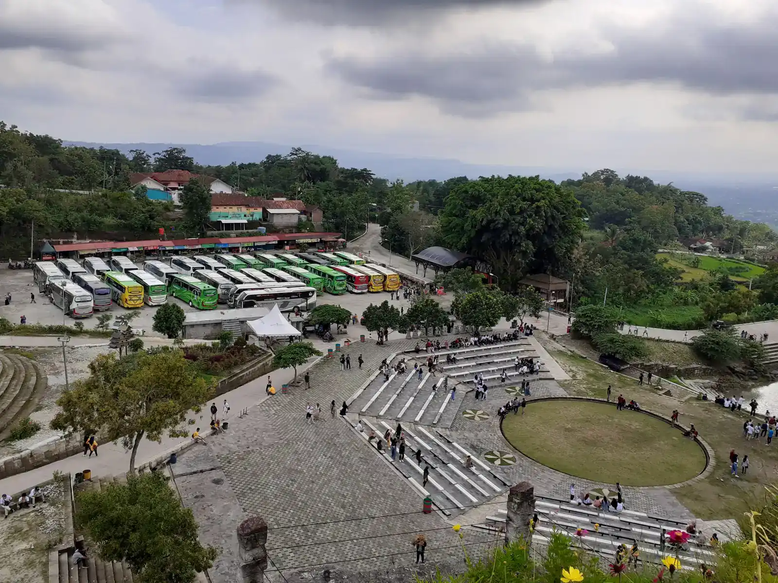 Amfiteater terbuka dengan tribun bertingkat dan panggung rumput bundar, di belakangnya parkiran bus wisata di Tebing Breksi