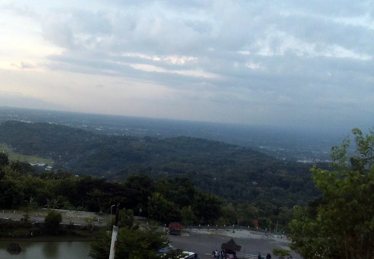 Pemandangan perbukitan hijau dan dataran Sleman di bawah langit mendung dilihat dari atas Tebing Breksi