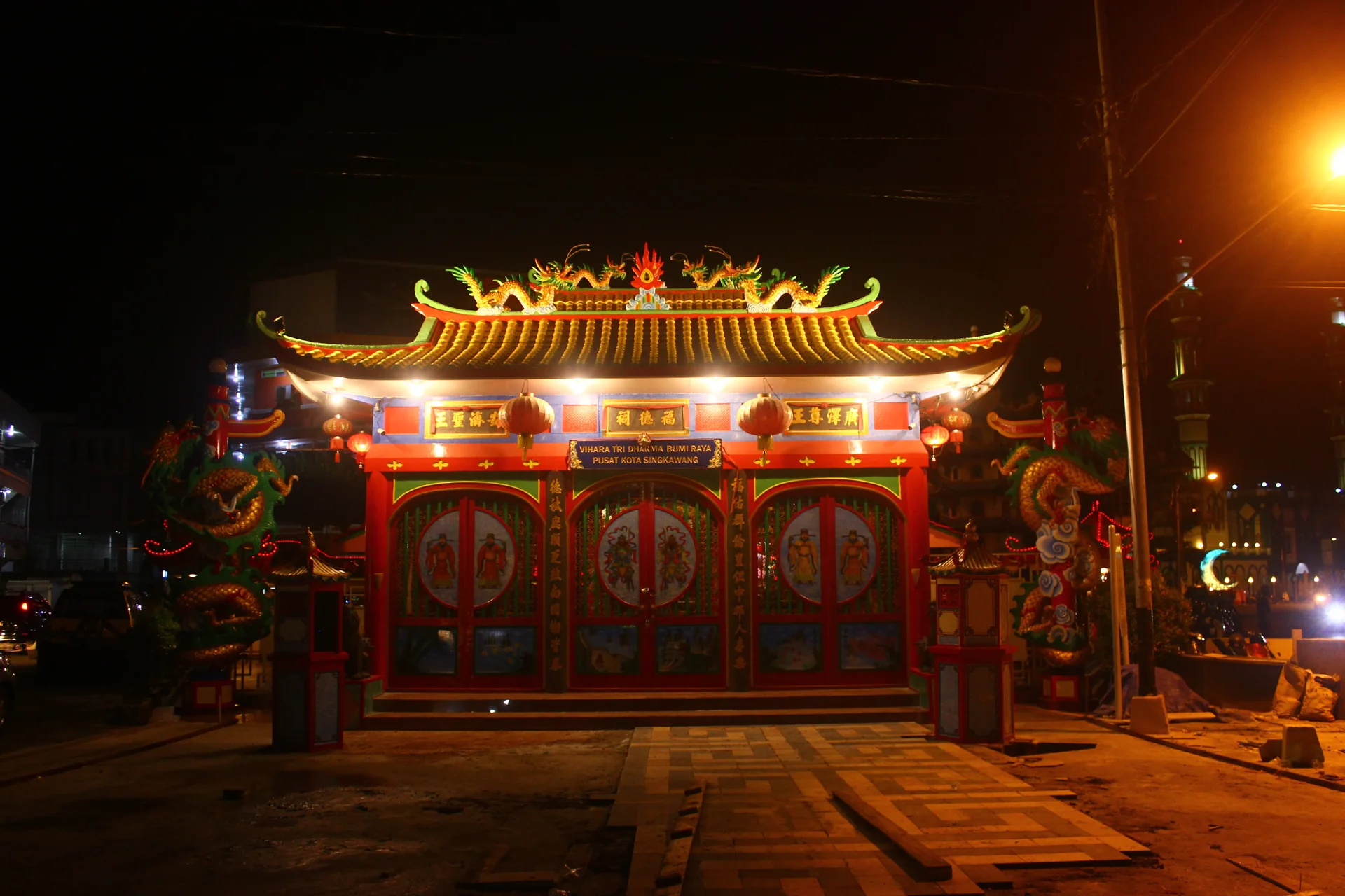 Fasad Vihara Tri Dharma Bumi Raya di Singkawang pada malam hari, dengan atap berhias naga, pintu merah bergambar dewa penjaga, dan lampion kecil
